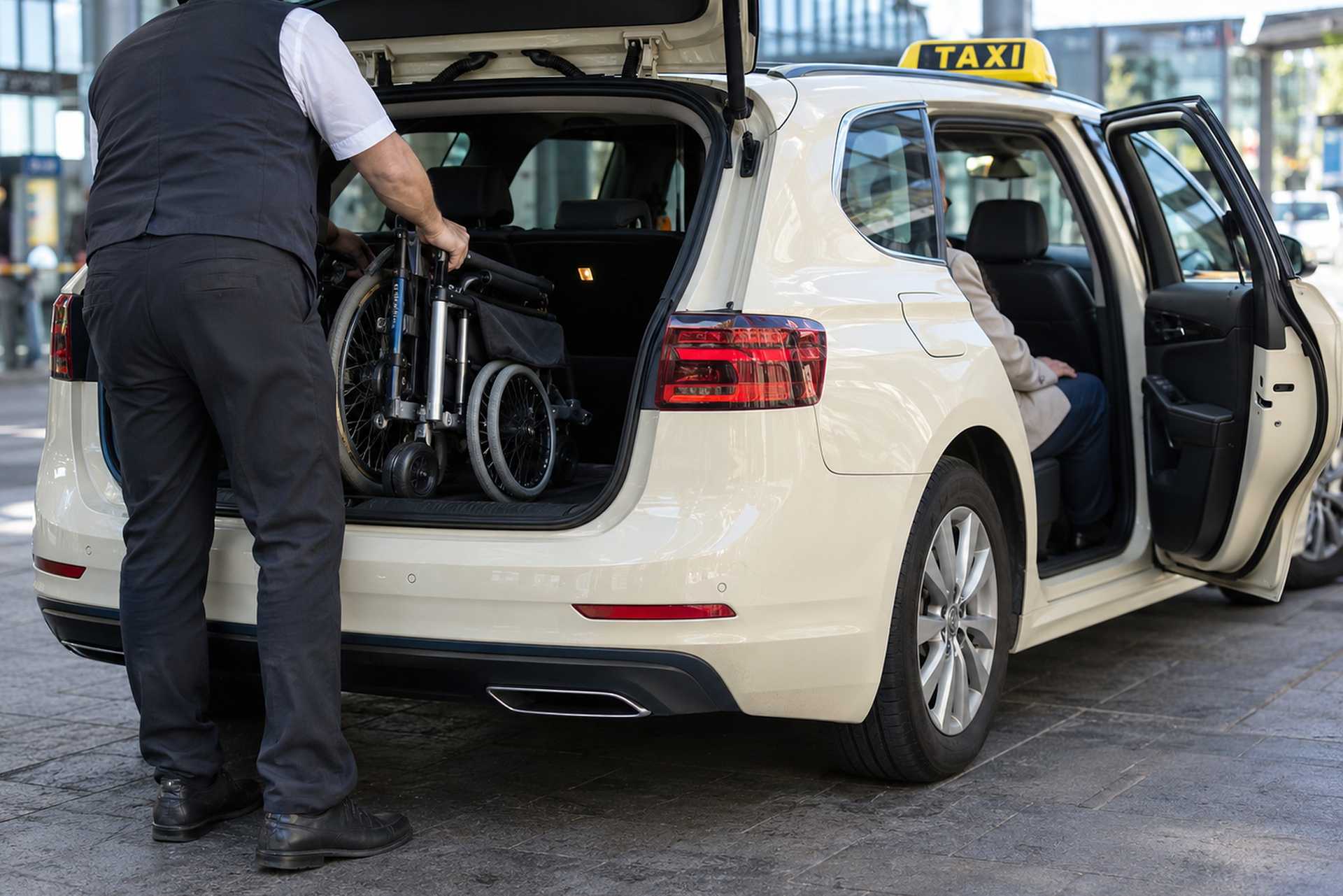 Symbolbild: Taxifahrer verlädt einen Faltrollstuhl in den Kofferraum, der Fahrgast sitzt auf dem Rücksitz